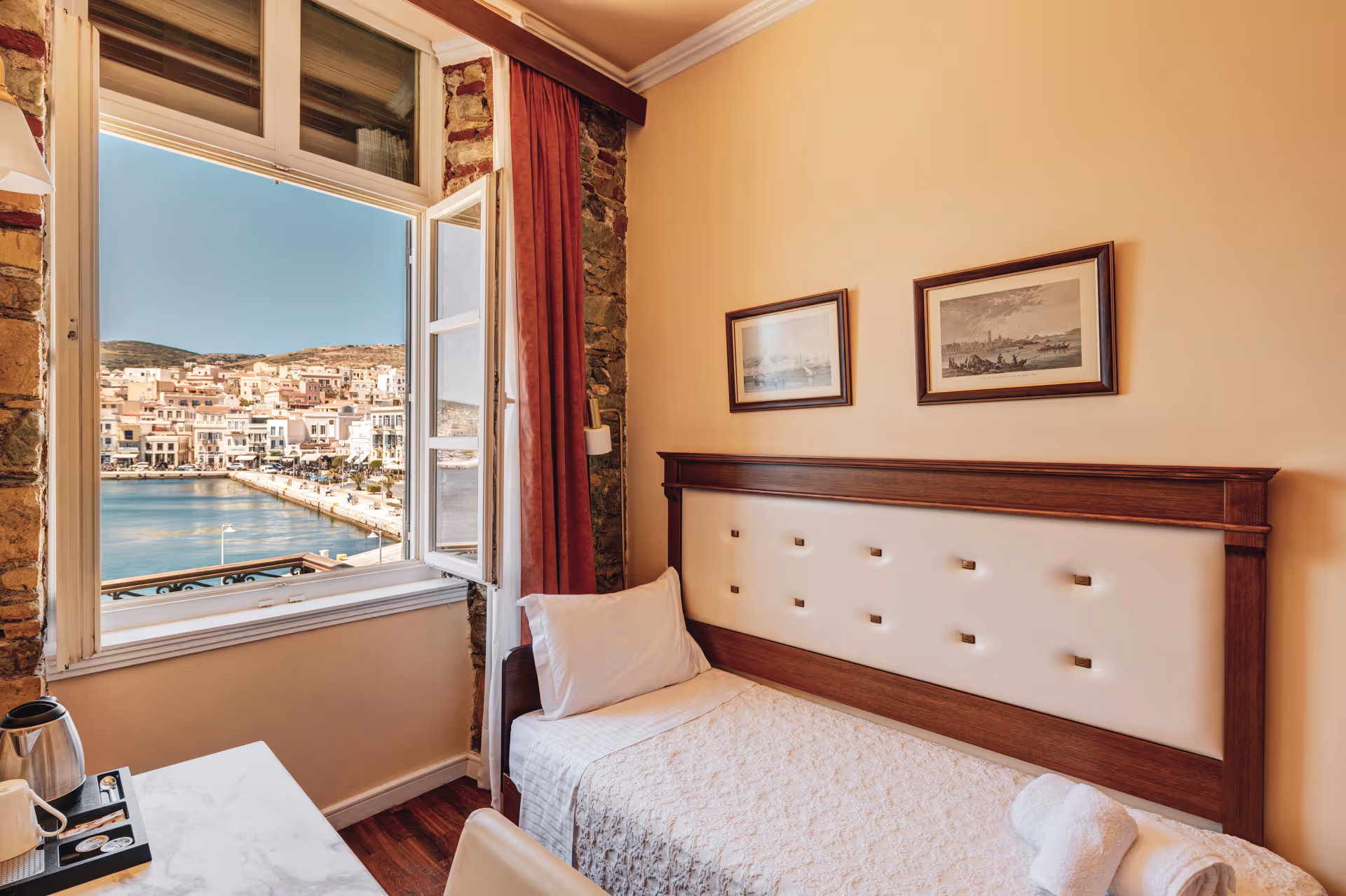 Cozy single bed with white linens and wooden headboard next to an open window showing a waterfront town view.