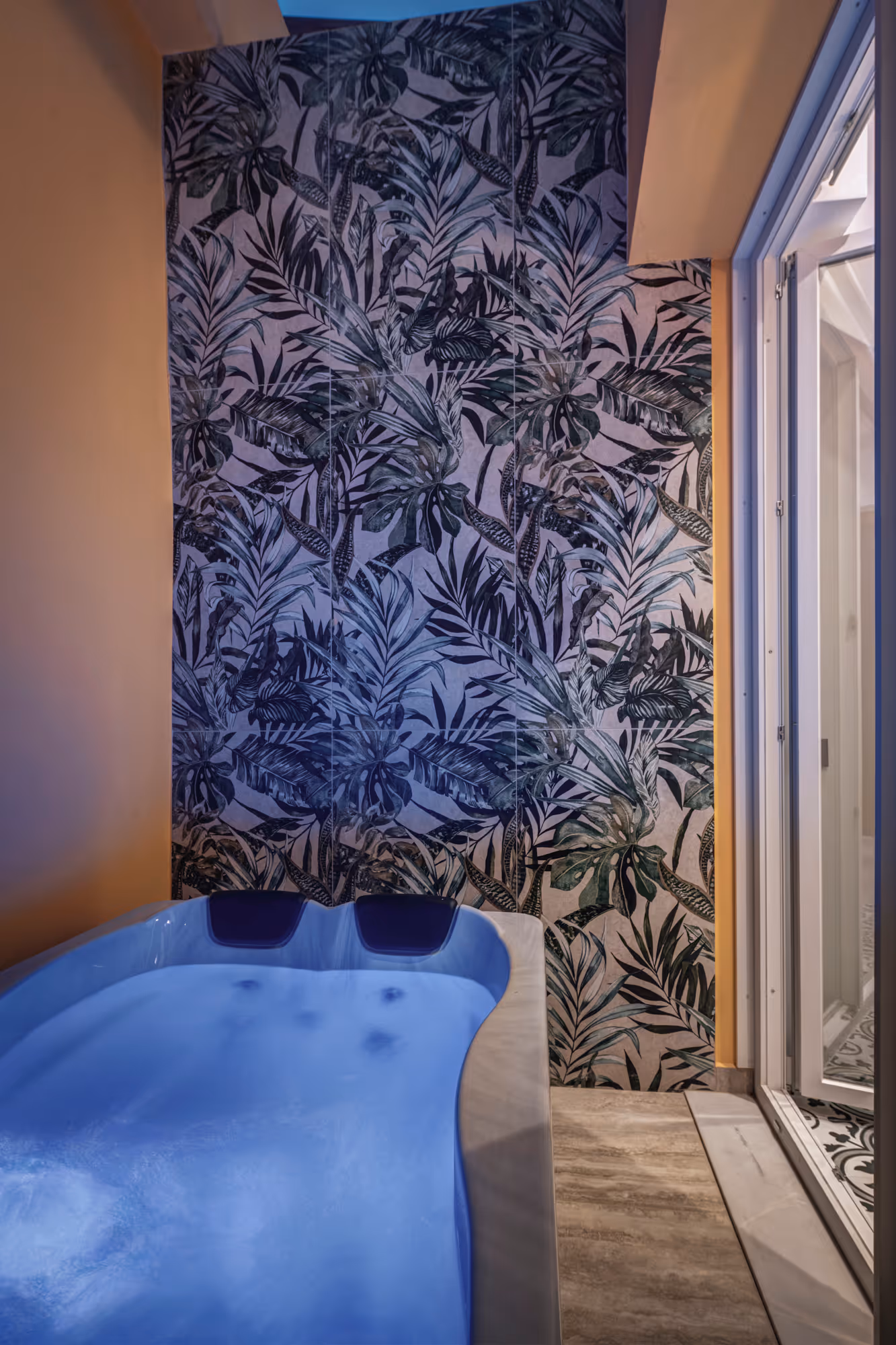 Indoor hot tub with blue water and two headrests, next to a tropical leaf patterned wall and glass door.