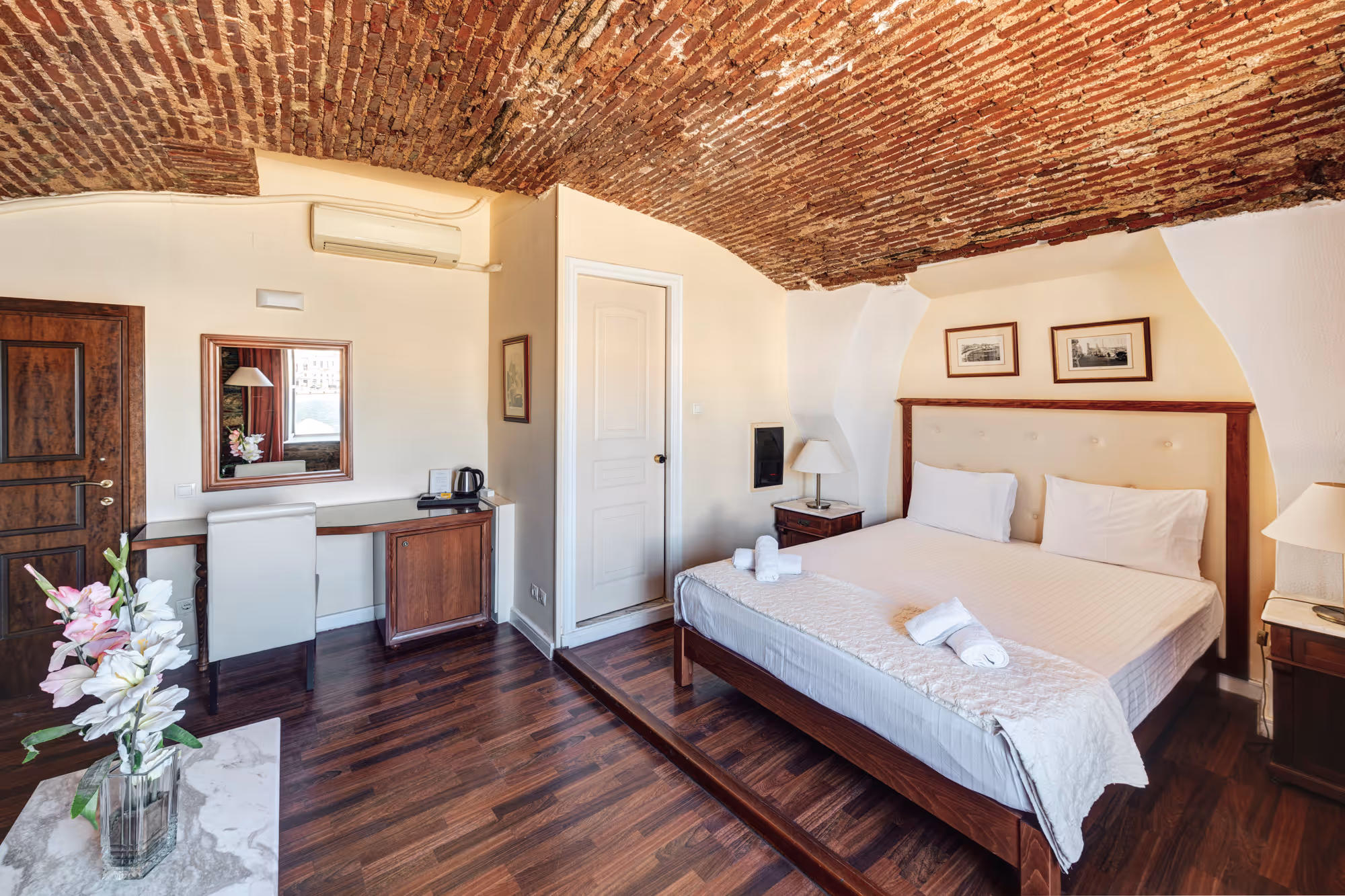 Hotel room with a large bed, exposed brick vaulted ceiling, wooden flooring, white walls, and a small desk with a mirror and chair.