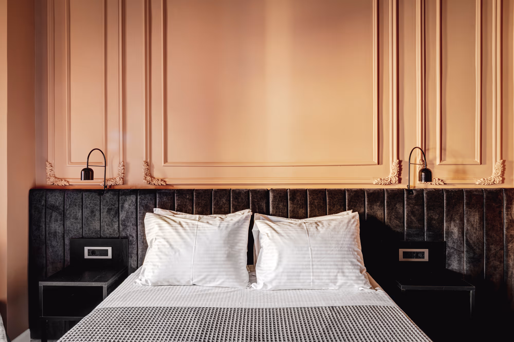 Modern bed with white pillows, gray textured blanket, dark tufted headboard, and two black wall lamps on beige paneled wall.